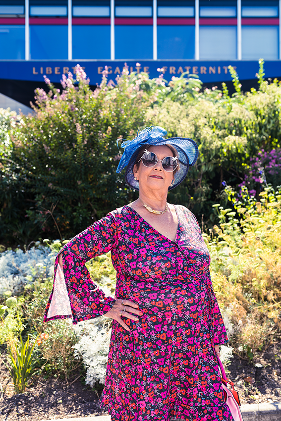 Chapeau bleu, lunette de soleil, robe fleurie et colorée, une personne âgée pose tout sourire devant la mairie. - 6 