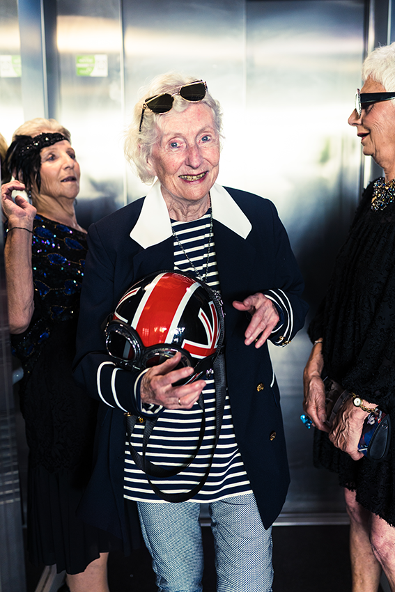 Lunettes de soleil sur la tête, une personne âgée pose tout sourire avec son casque de vélo aux couleurs de la Grande Bretagne à la main dans l'ascenseur de la Mairie. A l'arrière plan, deux de ses collègues se regardent amusées. - 9 
