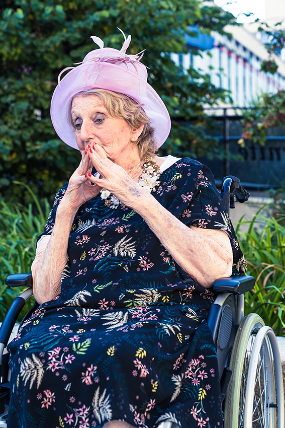 Personne âgée pensive posant en fauteuil roulant et vêtu d'un chapeau élégant couleur lilas devant le parterre fleuri de la mairie - 5 