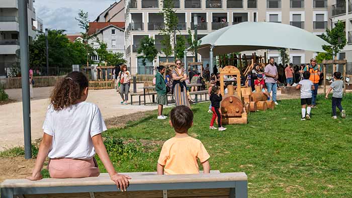 Au premier plan, deux enfants installés sur un grand transat en bois. Au second plan, l'aire de jeux ombragée investis par de nombreux enfants et parents.