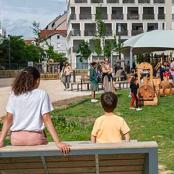 Au premier plan, deux enfants installés sur un transat en bois. Au second plan, l'aire de jeux ombragée investie par de nombreux enfants et parents.
